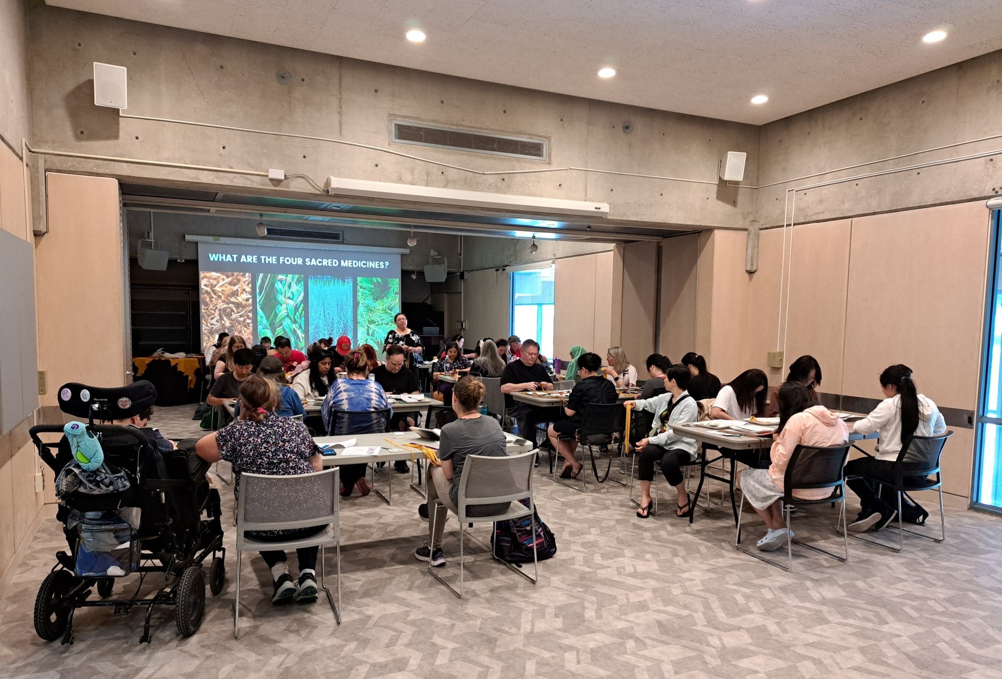Wide-angle view of workshop participants seated at tables crafting smudge feathers while following an Indigenous cultural presentation on screen during a Miskwaadesi Studio workshop in Richmond Hill, Ontario.